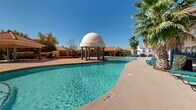This expansive outdoor pool area showcases a well-appointed resort-style retreat designed in a Spanish Colonial aesthetic. The centerpiece of the space is a generously sized, freeform swimming pool with crystal-clear turquoise water, accessible via multiple entry points including stainless steel handrails and steps. A striking architectural focal point stands poolside — a columned pavilion topped with a smooth, dome-shaped roof, adding a distinctive and elegant character to the environment.

The surrounding deck is finished in a neutral concrete surface, providing ample space for lounging and circulation. Rows of teal-colored chaise lounge chairs are arranged around the pool perimeter, offering plentiful seating for sunbathers. Shaded seating areas with teal market umbrellas are positioned throughout the space, providing relief from direct sun exposure.

The pool area is framed by mature fan palms that lend a tropical atmosphere, while a clubhouse-style building with a terracotta tile roof and a covered veranda borders one side, offering additional shade and sheltered seating. Wrought iron fencing defines the boundary of the space, and the entire area benefits from abundant natural sunlight under clear, open skies, creating a bright and inviting outdoor amenity.