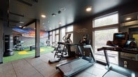 Located at 3110 Kinsrow Ave in Eugene, Oregon, this well-appointed fitness center is designed to support a full range of training activities within a single, open-plan floor. The space is divided into distinct functional zones, each purpose-built to accommodate different workout styles. The cardio area features multiple treadmills, elliptical trainers, a stationary bike, and a rowing machine, all positioned along expansive windows that frame views of the surrounding neighborhood and flood the space with natural light. A dedicated strength and weight training section is equipped with resistance machines, free weights, and a cable system, complemented by wall-mounted mirrors that enhance the sense of openness. The standout feature is a vibrant green and yellow artificial turf zone, evoking a sports field aesthetic and providing space for functional training, agility drills, and dynamic movement exercises. A freestanding boxing dummy adds a combat training element to this area. The interior design is defined by a bold dark color palette — deep charcoal walls and ceilings paired with rubber tile flooring — creating a focused, high-energy atmosphere. Motivational wall text and sports-themed mural artwork reinforce the performance-driven character of the facility. Recessed lighting throughout ensures consistent illumination across all training zones.