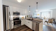 This thoughtfully designed single-floor residence at Greybarn Amityville offers a seamless blend of modern aesthetics and functional living. The open-concept kitchen is a standout feature, outfitted with crisp white shaker cabinetry, dark countertops, a classic white subway tile backsplash, and a full suite of stainless steel appliances including a refrigerator, range, microwave, and dishwasher. A peninsula with bar seating provides additional space for casual dining and entertaining. Rich hardwood flooring flows throughout the main living areas, anchoring the bright and airy atmosphere. The living room features vaulted ceiling details and transitions naturally into a dedicated dining nook framed by large windows that invite generous natural light and views of the surrounding community. The bedrooms are calm and well-proportioned, each with plush carpeting, ample closet space, and windows that continue the theme of natural illumination. The bathroom is finished in white subway tile extending from floor to ceiling, complemented by a wide vanity with dark countertops, a large mirror, and a full soaking tub with shower. Throughout the residence, the design strikes a balance between clean, contemporary finishes and a warm, welcoming ambiance.