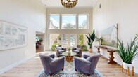 This well-appointed two-floor space offers a generous and versatile layout across two distinct levels. The main floor, the larger of the two, makes an immediate impression with its dramatic double-height ceilings in the primary living area. Floor-to-ceiling windows and French doors flood the space with natural light while framing lush outdoor views of surrounding greenery and a pool area. Light hardwood flooring runs throughout, complementing the neutral, warm-toned walls. A stylish chandelier anchors the seating area, which opens toward an entry foyer featuring double glass doors, additional tall windows, and display boards — suggesting a welcoming, community-oriented purpose. An adjacent dining and living room combination provides a more intimate gathering area, while a separate room on this level accommodates a conference-style setup with a large table, a private desk area, and direct exterior access. A compact workstation nook with wall-mounted monitor offers a focused area for individual use. The upper floor, accessed via a staircase, features a bright secondary living area with clerestory windows that invite natural light and outdoor tree views. Carpeted flooring on this level adds warmth and acoustic comfort. Together, both floors create a functional, elegantly styled environment suited for a variety of uses.