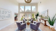 This well-appointed two-floor space offers a generous and versatile layout across two distinct levels. The main floor, the larger of the two, makes an immediate impression with its dramatic double-height ceilings in the primary living area. Floor-to-ceiling windows and French doors flood the space with natural light while framing lush outdoor views of surrounding greenery and a pool area. Light hardwood flooring runs throughout, complementing the neutral, warm-toned walls. A stylish chandelier anchors the seating area, which opens toward an entry foyer featuring double glass doors, additional tall windows, and display boards — suggesting a welcoming, community-oriented purpose. An adjacent dining and living room combination provides a more intimate gathering area, while a separate room on this level accommodates a conference-style setup with a large table, a private desk area, and direct exterior access. A compact workstation nook with wall-mounted monitor offers a focused area for individual use. The upper floor, accessed via a staircase, features a bright secondary living area with clerestory windows that invite natural light and outdoor tree views. Carpeted flooring on this level adds warmth and acoustic comfort. Together, both floors create a functional, elegantly styled environment suited for a variety of uses.