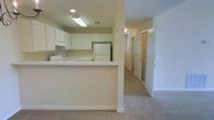 This single-floor space is designed with a practical and clean aesthetic, highlighted by neutral tones and a cohesive layout. The kitchen serves as a central feature, showcasing white upper and lower cabinetry that provides ample storage. A peninsula-style breakfast bar extends outward, offering additional counter space and a casual dining or workspace area. White appliances, including a refrigerator and range, complement the bright, uniform color scheme. Overhead, flush-mounted and fluorescent lighting ensures the kitchen remains well-lit.

Adjacent to the kitchen, the space transitions into a carpeted hallway that connects to additional rooms. The hallway reveals what appears to be a bathroom with a sliding glass shower door, as well as a closet with bi-fold doors, suggesting practical storage solutions throughout. The warm beige carpeting flows seamlessly from the main living areas into the corridor, contributing to a cohesive and comfortable atmosphere. The overall design is straightforward and functional, with white trim, neutral wall tones, and recessed lighting reinforcing a clean, move-in-ready environment suitable for everyday living.
