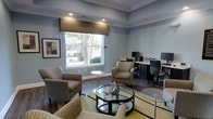 This single-floor space functions as a combined lounge and business center, thoughtfully designed to accommodate both relaxation and productivity. The room features soft blue-gray walls accented by clean white crown molding and a subtly raised ceiling detail, lending the space a refined, polished character. Recessed lighting provides even illumination throughout, complementing the natural light that filters in through a large window dressed with a structured, patterned valance. The window offers an outward view of mature trees and greenery, enhancing the calm atmosphere. The lounge area is furnished with upholstered armchairs in neutral tones, arranged around a round glass-top coffee table set atop a geometric yellow-and-white area rug, adding a warm contrast to the cool wall tones. Dark hardwood-style flooring runs throughout, grounding the space with a sense of warmth and cohesion. Along the far wall, a dedicated business center provides computer workstations with ergonomic seating, catering to work or study needs. Framed artwork on the walls adds a finishing decorative touch. Overall, the space balances functional amenities with comfortable, contemporary design.