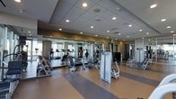 This fitness center offers a thoughtfully designed workout environment across a single level, providing ample space for a diverse range of exercise activities. The layout is open and well-organized, allowing for comfortable movement between equipment stations without congestion.

The strength training area features a multi-station cable machine system alongside a full dumbbell rack, offering options for both isolated and compound exercises. Multiple adjustable benches are positioned throughout the space to complement the free weights and resistance equipment. The cardio section includes treadmills and additional aerobic machines, catering to users with varying fitness goals.

The flooring consists of a durable, cushioned surface with a striped design in neutral tones, providing both safety and comfort underfoot. A large mirrored wall runs along one side of the room, enhancing the sense of openness while allowing users to monitor their form during workouts.

Recessed lighting is embedded in a drop-panel ceiling, delivering consistent, bright illumination throughout the space. Windows along the perimeter bring in natural light and contribute to an energizing atmosphere. The overall ambiance is clean, modern, and functional, reflecting a facility designed to accommodate a broad range of fitness needs efficiently.