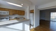 This single-story space centers around an open-concept kitchen that flows naturally into the adjacent living areas. The kitchen is outfitted with warm honey-toned wood cabinetry offering generous storage across both upper and lower units. A white refrigerator and dishwasher provide full functionality, while a single-basin sink with a standard faucet is positioned along the countertop. A peninsula-style breakfast bar extends outward, creating a natural division between the kitchen and the surrounding space, and offering additional surface area for casual dining or meal preparation. Recessed and fluorescent lighting illuminate the workspace effectively. The flooring transitions to warm hardwood throughout the adjoining hallway and living areas, adding a cohesive, natural element to the interior. White walls and white trim maintain a clean, neutral aesthetic throughout. A hallway visible from the kitchen leads toward additional rooms, including what appears to be a closet with louvered doors. Natural light filters into the space, enhancing the open and airy feel of the layout. The overall design is practical and straightforward, with a functional floor plan suited for everyday living.