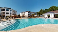 Located at 4645 Gordon Rd in Wilmington, North Carolina, this outdoor amenity space is designed to deliver a resort-style experience for its residents. The centerpiece is a generously sized swimming pool with crystal-clear water, framed by a wide concrete pool deck lined with lounge chairs and shaded umbrellas, offering ample space for relaxation under the Carolina sun.

Adjacent to the pool is a thoughtfully designed covered cabana pavilion, distinguished by its vaulted wood-paneled ceiling and white-painted exterior with brick-accented column bases that add a charming, coastal aesthetic. The structure opens on multiple sides to capture natural light and refreshing views of the surrounding greenery and pool area. Inside the cabana, comfortable sectional and accent seating is arranged around a wood coffee table atop a patterned area rug, creating an inviting lounge atmosphere. A wall-mounted fireplace and flat-screen television enhance the space for year-round enjoyment.

The pavilion's open-air design flows seamlessly to an adjacent patio area furnished with additional seating, vibrant potted plants, and wrought-iron railings overlooking mature trees. The multi-story residential buildings, featuring white facades with dark-trimmed windows, provide a polished architectural backdrop, while palm trees and native landscaping reinforce the warm, coastal character of this well-appointed outdoor retreat.
