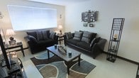 This single-floor space includes a living room designed with a contemporary aesthetic and a coordinated color scheme. The living room centers around two large, dark upholstered sofas accented with patterned throw pillows in grey and black tones, arranged to encourage comfortable conversation. A dark wood coffee table with a marble-style surface sits at the heart of the seating arrangement, resting atop a geometric area rug in shades of teal, brown, and cream that adds visual interest to the light-toned flooring. Natural light enters the room through a window fitted with white horizontal blinds, brightening the neutral white walls. Decorative touches include a table lamp providing warm ambient lighting and a wall-mounted photo collage frame with ornamental iron detailing, adding a personalized element to the space. The overall ambiance is modern and cohesive, with the dark furniture contrasting effectively against the light walls and flooring to create a bold yet inviting atmosphere throughout the living area.