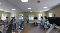 This single-floor fitness center offers a practical and well-organized workout environment suited for a range of exercise routines. The space is outfitted with a selection of strength training equipment, including a multi-station cable machine and an adjustable weight bench, alongside cardio machines such as an elliptical trainer and a stationary bike. The layout is thoughtfully arranged to allow comfortable movement between stations. A large floor-to-ceiling mirror spans one wall, visually expanding the space and allowing users to monitor their form during exercise. The room is finished with neutral olive-toned walls and carpeted flooring, contributing to a clean and understated aesthetic. Recessed panel lighting in the ceiling provides consistent, even illumination throughout. Multiple windows along the exterior wall invite natural light into the space and offer views of the surrounding greenery, creating an airy and pleasant atmosphere for workouts. The overall design is functional and accessible, making it a convenient facility for daily fitness use.