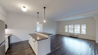 Located at Greybarn Amityville in Amityville, NY, this single-floor residence combines modern design with functional living. The open-concept kitchen and living area serves as the heart of the home, anchored by a central island with a dark quartz countertop, white shaker cabinetry with matte black hardware, and classic white subway tile backsplash. Pendant lighting hangs above the island, complementing the stainless steel appliances including an electric range and microwave. Rich, dark hardwood flooring flows throughout the main living areas, creating a warm contrast against the crisp white walls. Large windows invite generous natural light into the expansive living room, which features exposed beam details and direct access to an entry area. The bedrooms are carpeted for comfort, each offering bright windows with outdoor views and individual climate control units. The bathrooms are elegantly appointed with floor-to-ceiling white subway tile, mosaic tile flooring, dark quartz vanity countertops, white shaker cabinetry, large mirrors, and a soaking tub with shower combination. An in-unit laundry area adds everyday convenience. The clean, neutral palette and thoughtful finishes throughout create a cohesive, contemporary living environment.