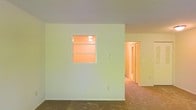 This single-floor space presents a straightforward layout with a calm, neutral aesthetic throughout. The main living area is finished with wall-to-wall carpeting in a muted tone, contributing to a quiet and understated atmosphere. A notable architectural detail is the interior pass-through window set into a dividing wall, framed cleanly and illuminated from the adjoining room, creating a visual connection between spaces while maintaining separation. The walls are painted in a soft, light hue that keeps the interior feeling open and airy. A hallway visible through an open doorway leads further into the unit, where folding closet doors can be seen, suggesting additional storage or bedroom access beyond. The layout appears to flow from the main living space into a corridor connecting the remaining rooms. Lighting is modest, with a ceiling-mounted fixture visible through the pass-through window providing warm illumination. The overall design is functional and clean, with minimal ornamentation, making it a versatile canvas suitable for a variety of uses and furnishing styles.