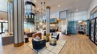 Located at 1260 NE Reserve Trail in Jensen Beach, Florida, this single-story clubhouse delivers a sophisticated, resort-inspired atmosphere through thoughtful architectural design and curated interior details. The grand main lounge immediately commands attention with its soaring double-height ceilings, gold-accented structural columns, and a dramatic suspended fireplace encased in a geometric frame — serving as the visual centerpiece of the space. Light wood flooring runs throughout, complementing an eclectic mix of furnishings that include woven lounge chairs, leather ottomans, and richly patterned sofas arranged across multiple seating areas. Striking teal mosaic tile walls add bold color and texture, while an array of pendant lighting fixtures creates warm, layered illumination. A bar or lounge area with counter seating is visible toward the rear, offering an additional gathering point. A dedicated reading nook features floor-to-ceiling built-in shelving and a plush upholstered sofa, ideal for quiet relaxation. Floor-to-ceiling glass walls and double doors open toward an outdoor pool area, allowing abundant natural light to pour in while framing views of the resort-style pool and landscaping beyond. The entry foyer features wood-paneled walls and a slatted divider screen, establishing an elegant transition into the main social spaces.