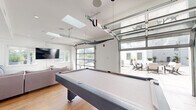 This contemporary single-floor space is thoughtfully designed for entertainment and social gatherings, combining modern aesthetics with versatile functionality. The centerpiece of the space is a spacious game room anchored by a full-sized pool table, flanked by comfortable sectional seating and a wall-mounted flat-screen television. A skylight overhead floods the area with natural light, while a full-panel glass garage door opens the room directly to an outdoor patio, creating a seamless indoor-outdoor flow and expanding the entertainment area considerably. The lounge zone features light hardwood flooring throughout, clean white walls with bold navy accent walls, and recessed lighting that complements the abundant natural light.

The kitchen is sleek and modern, designed in a galley-style layout with crisp white cabinetry, quartz countertops, and a geometric hexagonal tile backsplash. Stainless steel appliances, including a refrigerator, range, and dishwasher, provide full functionality. A kitchen island with bar seating adds additional prep and dining space. Adjacent to the kitchen, a dining area with bar-height seating offers another gathering spot. Throughout the space, the consistent use of light wood flooring, white tones, and minimalist finishes creates a bright, clean, and cohesive atmosphere ideal for hosting.