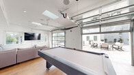 This contemporary single-floor space is thoughtfully designed for entertainment and social gatherings, combining modern aesthetics with versatile functionality. The centerpiece of the space is a spacious game room anchored by a full-sized pool table, flanked by comfortable sectional seating and a wall-mounted flat-screen television. A skylight overhead floods the area with natural light, while a full-panel glass garage door opens the room directly to an outdoor patio, creating a seamless indoor-outdoor flow and expanding the entertainment area considerably. The lounge zone features light hardwood flooring throughout, clean white walls with bold navy accent walls, and recessed lighting that complements the abundant natural light.

The kitchen is sleek and modern, designed in a galley-style layout with crisp white cabinetry, quartz countertops, and a geometric hexagonal tile backsplash. Stainless steel appliances, including a refrigerator, range, and dishwasher, provide full functionality. A kitchen island with bar seating adds additional prep and dining space. Adjacent to the kitchen, a dining area with bar-height seating offers another gathering spot. Throughout the space, the consistent use of light wood flooring, white tones, and minimalist finishes creates a bright, clean, and cohesive atmosphere ideal for hosting.