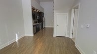 This two-storey space showcases a thoughtfully designed open-concept layout, where the main living area flows seamlessly into a well-appointed kitchen. The flooring throughout is a warm, medium-toned hardwood-style material that adds continuity and a grounded aesthetic across the main level.

The kitchen is tucked into a defined nook and features dark cabinetry with recessed lighting overhead, offering a striking contrast against the neutral grey walls. Stainless steel appliances, including a refrigerator, stove, and dishwasher, are integrated into the design, giving the kitchen a sleek, functional character.

The main living area benefits from soaring ceilings that extend upward toward the second floor, creating a strong sense of vertical space and openness. The neutral grey walls and white trim provide a clean, versatile backdrop throughout.

A white interior door leads to additional areas of the unit, while what appears to be a mirrored surface or glass panel is visible along one wall, adding depth and light to the space. The overall ambiance is modern and understated, with a practical yet polished finish throughout.