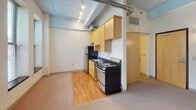 Located on S Eighth St in the heart of downtown Minneapolis, this single-floor unit showcases an industrial loft aesthetic defined by exposed concrete ceilings with visible structural beams and overhead ductwork running throughout. Track and fluorescent lighting complement the raw architectural character overhead.

The open main living area is generously proportioned, featuring neutral carpet flooring and cream-toned walls that create a bright, airy atmosphere. A bank of tall windows lines the living space, allowing natural light to filter in while framing views of the surrounding urban streetscape. A separate bedroom offers similar exposed ceiling details, carpeted flooring, and additional windows with city views, providing a quiet, well-lit retreat.

The kitchen is efficiently designed with light wood cabinetry, dark countertops, hardwood flooring, and stainless appliances including a range and refrigerator. Exposed ductwork continues into this space, reinforcing the loft-style character. A sliding barn-style door leads toward the bathroom.

The bathroom is cleanly appointed with a pedestal sink, round mirror, a full tub-shower combination, and classic penny tile flooring, offering a functional and tidy finish to the space. Overall, the unit delivers an authentic urban loft experience in a prime downtown Minneapolis location.