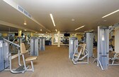 This fitness center offers a comprehensive workout environment spread across a single, open floor plan. The space is thoughtfully organized into distinct activity zones, providing a seamless flow between cardio, strength, and functional training areas.

The cardio section features a row of treadmills positioned along large windows that invite natural light and offer outdoor views, creating an energizing atmosphere for endurance workouts. Nearby, a selection of upright and recumbent stationary bikes, ellipticals, and other cardio equipment round out this zone.

The strength training area is anchored by a well-stocked dumbbell rack with a flat bench for free weight exercises, accompanied by a ceiling-mounted flat-screen television. Full-length mirrors line the walls, visually expanding the space and allowing users to monitor their form. Stacked exercise mats are readily available for floor-based workouts and stretching.

Dedicated selectorized machines — including leg press and cable stations — are positioned throughout the floor, supported by sturdy white columns that define the layout. The flooring throughout combines durable carpet tile with hard surface sections suited for various exercise modalities.

Lighting is provided by sleek linear LED fixtures set into a neutral-toned ceiling, maintaining a bright and focused environment. The overall ambiance is clean, modern, and functional, making this fitness center well-suited for a wide range of fitness goals.