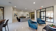 This contemporary single-floor communal space is designed with a clean, modern aesthetic that blends functionality with sophisticated style. The main lounge area features polished light-toned tile flooring that reflects recessed ceiling lighting, creating a bright and welcoming atmosphere. Comfortable seating — including a channeled gray sofa and a pair of bold blue velvet accent chairs with gold legs — is thoughtfully arranged to encourage relaxed gathering, while colorful abstract artwork adorns the crisp white walls, adding a vibrant artistic touch.

Large floor-to-ceiling glass partitions with sleek black metal framing divide the space into distinct functional zones while maintaining a sense of openness and visual connectivity. One glass-enclosed room serves as a private meeting or conference area, outfitted with seating and visible artwork. An adjacent lounge corridor lined with the same black-framed glass panels leads further into the space, offering views into a well-lit interior common area.

A dedicated dining or collaboration nook features a substantial dark wood high-top table with dark upholstered chairs, positioned beside oversized windows that frame views of the surrounding building complex and outdoor amenities. Natural light pours in generously through these large windows, enhancing the airy, open feel of the entire space. The overall design is cohesive, professional, and inviting — well-suited for both social interaction and productive use.