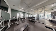This fitness center is a thoughtfully designed, single-floor facility built to accommodate a full range of workout routines. The space is divided into distinct functional zones, seamlessly connected by durable rubber flooring throughout. The cardio area features a row of treadmills, elliptical machines, and a stair climber, all positioned near large windows that invite abundant natural light and views of the surrounding exterior. A ceiling fan mounted within a tray ceiling overhead enhances air circulation and comfort during workouts. The strength training zone is well-appointed with a multi-station cable machine, a squat rack with a flat bench, and a tiered dumbbell rack stocked with a wide range of free weights. A floor-to-ceiling mirror wall visually expands the space and allows users to monitor their form. Additional equipment includes a rowing machine, further broadening the training options available. The walls feature a calming sage green accent color complemented by crisp white trim and recessed lighting, lending the space a clean, modern aesthetic. A wall-mounted television and an interior window overlooking an adjacent area add further functionality and openness to the overall environment.