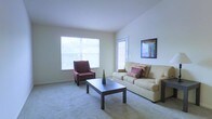 This single-floor space features a clean, straightforward layout centered around a comfortable living area. The living room is finished with wall-to-wall carpet in a neutral tone, contributing to a warm and understated aesthetic throughout. Walls are painted in a light, off-white hue that enhances the sense of openness and brightness. A large window along one wall allows generous natural light to fill the room, creating an airy atmosphere. An additional door with a sidelight panel provides both access and supplementary light to the space. The vaulted ceiling adds vertical dimension, making the room feel more spacious than its footprint might suggest. Furnishings in the image include a beige sofa accented with decorative pillows, a deep red accent chair, and a dark rectangular coffee table, illustrating the room's capacity for comfortable, functional seating arrangements. A framed piece of wall art adds a subtle decorative touch to the otherwise minimal interior. The overall ambiance is calm and neutral, offering a versatile foundation that can easily be adapted to a variety of interior styles and personal preferences.