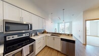 This thoughtfully designed single-floor residence combines contemporary style with everyday functionality. The kitchen is a standout feature, outfitted with crisp white shaker cabinetry, light quartz countertops, a gray subway tile backsplash, and a full suite of stainless steel appliances including a range, microwave, and dishwasher. Pendant lighting with Edison-style bulbs adds warmth, while a window above the sink brings in natural light and outdoor views.

The kitchen flows seamlessly into the open living area via a peninsula breakfast bar with seating — an ideal setup for casual dining or entertaining. The living space features warm wood-tone flooring and track lighting overhead, creating an inviting and versatile environment.

The bedroom is generously proportioned and bathed in natural light from expansive floor-to-ceiling windows that offer pleasant outdoor views. Soft neutral carpeting adds comfort underfoot.

The bathroom is finished to a high standard, featuring a wide vanity with quartz countertops, an undermount sink, modern fixtures, and a large mirror with overhead lighting. Warm wood-tone cabinetry and doors complement the clean, contemporary aesthetic throughout.

Overall, the space presents a cohesive modern design with an abundance of natural light, quality materials, and a practical, open layout.