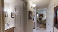 This single-story space presents a thoughtfully designed interior that connects its living areas through a central hallway. The corridor is characterized by textured walls in a neutral tone, accented by an elegant wall-mounted sconce that provides warm, ambient lighting. A classic six-panel door adds a traditional architectural touch near the entryway.

The hallway serves as a natural transition point, offering glimpses into the adjoining spaces. White cabinetry is visible toward the kitchen area, suggesting a clean and functional layout. The living area beyond features light-toned carpeting and is furnished with classic seating, including an upholstered armchair and ottoman, creating a comfortable and inviting atmosphere.

Wall art in dark wood frames is thoughtfully placed throughout, contributing to a warm, homey aesthetic. Crown molding details are visible in the living area, adding a refined architectural element. The overall color palette leans toward soft neutrals — creamy whites, warm beiges, and light taupes — creating a cohesive and calming environment throughout the space. Natural light filters in from windows in the living area, further enhancing the open and airy feel of the interior.