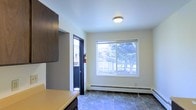 This single-floor space presents a clean and functional interior with a straightforward layout. The main area visible features stone tile flooring, which adds texture and durability underfoot, complementing the neutral, light-colored walls throughout. A large sliding window dominates one wall, allowing natural light to fill the space while offering a pleasant view of surrounding trees and neighboring structures outdoors, contributing to an open and airy feel.

A flush-mounted ceiling light provides additional illumination, keeping the space well-lit. A baseboard heating unit runs along the lower portion of the exterior wall beneath the window, offering an efficient heating solution. Dark-framed doorways provide strong visual contrast against the light walls, lending a degree of architectural definition to the space.

Dark cabinetry is partially visible along one side of the room, suggesting integrated storage or kitchen elements nearby. A fire extinguisher is mounted near the entry door, indicating attention to safety. The entry door, framed in dark trim and featuring a glass panel, provides access to the outdoors and reinforces the connection between the interior and exterior environment. Overall, the space is practical, tidy, and well-maintained.