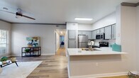 This single-floor residence offers a thoughtfully designed open-concept layout that seamlessly connects the kitchen, dining, and living areas. The kitchen is a standout feature, showcasing gray shaker-style cabinetry, a classic white subway tile backsplash, stainless steel appliances including a range, microwave, and refrigerator, and a white quartz peninsula with an undermount sink that doubles as a breakfast bar. Light wood-style flooring runs throughout the main living areas, enhancing the bright and airy atmosphere. The living room is bathed in natural light from a sliding glass door that opens to the outdoors, and is accented with crown molding for an elevated finish. A dedicated dining area sits adjacent to the kitchen, providing a practical and inviting space for meals. The primary bedroom is a serene retreat with soft carpeting, a ceiling fan, natural light from large windows, and a private en-suite bathroom featuring a quartz vanity and a tub-shower combination. A second bedroom, equally well-lit and carpeted, includes access to a walk-in closet. The second full bathroom mirrors the same clean, modern aesthetic with gray cabinetry, a white quartz vanity, and a tiled tub-shower. A long hallway connects all private spaces, contributing to a functional and comfortable floor plan.