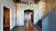 Located within the historic Germantown Mill Lofts, this three-floor residence is a compelling example of industrial adaptive reuse architecture. The space preserves the building's storied past through its raw, textured exposed brick walls, which display layers of aged paint that add depth and authenticity to the interior. A dramatic arched brick doorway serves as a defining architectural centerpiece, framing transitions between living areas with historic elegance.

Rich, dark hardwood floors run throughout, grounding the space and contrasting beautifully with the weathered masonry walls. Overhead, exposed ductwork and bold red piping trace the ceiling, reinforcing the loft's industrial aesthetic while adding visual interest. Pendant lighting fixtures hang from above, casting warm, focused illumination that complements the space's moody, textured ambiance.

The open layout allows for a generous flow between areas, with a kitchen island visible through the arched passage, hinting at a practical yet characterful culinary space. A staircase connects the multiple levels, offering a sense of vertical scale. The overall atmosphere is one of urban sophistication, blending historic industrial heritage with modern livability.