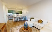 This thoughtfully designed 600-square-foot, one-bedroom, two-bathroom apartment combines modern aesthetics with functional living. The open-concept kitchen and living area serves as the heart of the unit, anchored by a striking kitchen outfitted with sage green shaker cabinets, granite countertops, a white subway tile backsplash, stainless steel appliances, and brass hardware. A large island with seating for four sits beneath a pair of gold geometric pendant lights, creating an ideal space for dining and entertaining. The living room features warm hardwood-style flooring, neutral walls, and large windows that invite abundant natural light. The primary bedroom measures 140 square feet and includes a decorative wall panel accent, windows with exterior views, and direct access to the en-suite bathroom. The en-suite bath showcases a green vanity with granite countertops, brass fixtures, a framed mirror, and a glass-enclosed walk-in shower with subway tile surround. A walk-in closet with built-in rod storage and a stacked washer/dryer adds everyday convenience. A private patio overlooks a landscaped outdoor area with views of the surrounding community. The unit's cohesive design palette of warm neutrals, sage green, and brushed gold creates a polished, contemporary ambiance throughout.