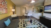 This single-floor residence in the United States features an open-concept design that seamlessly connects the living, dining, and kitchen areas into one cohesive space. The living area is anchored by a dark upholstered sofa and a glass coffee table, set atop a bold geometric-patterned area rug in teal, black, and white. Ceiling-mounted track lighting and a ceiling fan with light fixture provide ample illumination throughout the space.

The dining area, positioned centrally, includes a dark table surrounded by modern white chairs with chrome accents, creating a clean, contemporary aesthetic. Adjacent to this, an entryway features a coat rack and closet, offering practical storage near the front door.

The kitchen, visible from the living space, is separated by a raised breakfast bar that provides additional seating and countertop space. A sink is visible at the counter's edge, and a large mirror on the kitchen wall enhances the sense of openness. Accent walls painted in warm gold tones complement the neutral surrounding walls, adding depth and warmth to the overall ambiance of the space.
