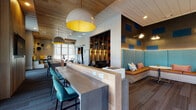 Located at 330 Pennington Centre Blvd in Nashville, TN, this single-floor community space is a thoughtfully designed hub for work, relaxation, and social connection. The interior showcases a contemporary aesthetic defined by warm wood-plank ceilings, textured carpeting, and a bold color palette of teal, burnt orange, and navy that carries throughout the space.

The central gathering area features a substantial concrete-topped communal bar with built-in power outlets and teal upholstered bar seating, illuminated by striking oversized pendant lights with vibrant yellow interiors. Adjacent to this is a cozy lounge nook outfitted with a tufted teal bench, a wall-mounted flat-screen TV, and a bold geometric accent wall in orange and navy, complemented by open wood shelving.

A bright, window-lined seating area provides an inviting retreat filled with natural light and outdoor views, furnished with comfortable chairs, a cushioned wraparound bench, and playful accent pillows. Sheer curtains soften the abundant daylight pouring in.

The dedicated business center offers individual workstations with built-in desks, teal seating, and wall-mounted monitors, while private focus nooks feature reclaimed wood accent walls, live greenery, and individual desks with accessible power outlets. Curated artwork adds a polished, creative finish throughout the entire space.