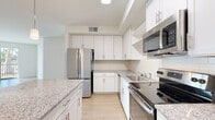 Located at 16520 San Carlos Blvd in Fort Myers, Florida, this single-floor residence offers a clean, contemporary design with a bright and open feel throughout. Light hardwood-style flooring flows seamlessly across the entire unit, complementing the neutral, warm-toned walls and white trim.

The kitchen is a standout feature, showcasing white shaker-style cabinetry, granite countertops, a subway tile backsplash, and a full suite of stainless steel appliances including a refrigerator, electric range, and microwave. A central island with pendant lighting above provides additional prep space and enhances the open-concept connection to the living area, which is generously proportioned and filled with natural light.

The bedrooms are well-sized, each featuring closets and windows with blinds that frame views of the surrounding Florida landscape, including glimpses of palm trees. Ceiling fans add to the comfort of the sleeping quarters.

The bathrooms are finished with granite-topped white vanities and hardwood-style flooring. The primary bathroom offers a dual-sink vanity with a large mirror and ample cabinet storage, while the secondary bathroom features a tub-shower combination. The overall ambiance of the space is fresh, modern, and move-in ready, reflecting a cohesive design aesthetic throughout.