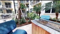 This expansive outdoor amenity area is designed to evoke a resort-like atmosphere, centered around a generously sized swimming pool with clear turquoise water. The pool deck is finished with elegant stone pavers and lined with a row of modern mesh lounge chairs, providing ample space for relaxation. Blue market umbrellas are positioned throughout the deck, offering shaded seating areas ideal for enjoying the outdoors. The courtyard setting is framed on multiple sides by a multi-story building featuring warm earth-toned stucco facades, large windows, and private balconies that overlook the amenity space below.

Mature palm trees are thoughtfully integrated throughout the landscape, contributing to the tropical ambiance and providing natural shade and visual interest. Lush, layered plantings of tropical foliage fill the garden beds, adding vibrant greenery that softens the hardscape elements. A standout feature of the space is a fire pit with a granite-topped surround and warm wood base, creating an inviting gathering area nestled within the landscaping. The combination of tropical greenery, resort-style amenities, and architectural detailing gives this outdoor space a polished, welcoming character.