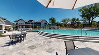 This single-level outdoor amenity space is part of a well-appointed residential community in the United States, designed to blend modern farmhouse architecture with functional, resort-style leisure. The centerpiece of the space is a generously sized swimming pool with a multi-step entry, framed by an expansive travertine tile deck that provides ample room for outdoor seating, dining, and recreational activities such as cornhole. Lounge chairs, umbrella-shaded tables, and casual seating arrangements are distributed throughout the deck, creating a welcoming environment for relaxation and social gatherings. A covered porch runs along the adjacent building, supported by stately white brick columns and featuring large, dark-framed floor-to-ceiling glass doors and windows that offer unobstructed views of the pool area. The porch provides a shaded transition zone between the interior and the sun-drenched pool deck, and is furnished with additional seating for outdoor enjoyment. The surrounding community showcases a mix of board-and-batten and traditional architectural styles with metal and pitched rooflines. Mature trees line the perimeter, adding natural shade and greenery under open, sunny skies — contributing to the bright, airy ambiance of this outdoor amenity hub.
