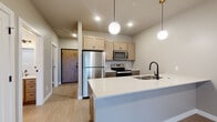 This single-floor unit presents a thoughtfully designed layout with modern finishes throughout. The open-concept kitchen serves as a central highlight, anchored by a generous quartz-topped peninsula with an undermount sink and matte black faucet. Light maple cabinetry lines the walls above stainless steel appliances, including a refrigerator, electric range, and microwave. Globe pendant lights hang overhead, adding a contemporary touch to the space. Light wood-tone luxury vinyl flooring runs consistently throughout the unit, contributing to a cohesive, airy aesthetic.

The living area is spacious and versatile, featuring recessed lighting, a wall-mounted TV bracket, and a built-in desk ledge along one wall. Sliding glass doors open directly to an outdoor balcony, allowing natural light to fill the room. An adjacent bedroom also benefits from balcony access and a large closet with a paneled door.

The bathroom is cleanly finished with warm wood-tone cabinetry, a white countertop, undermount sink, and matte black hardware accents, maintaining the modern design language found throughout. A dedicated laundry closet houses a full-size washer and dryer with wire shelving above, providing practical in-unit convenience. Overall, the unit offers a clean, contemporary living environment with functional amenities.