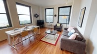 This well-appointed unit is spread across a single floor and offers a thoughtfully organized layout with multiple distinct living areas. The open living room serves as the heart of the space, featuring warm hardwood flooring, track lighting, and large black-framed windows that fill the room with natural light. Two gray sofas and an armchair create a comfortable seating arrangement centered around a modern coffee table, while a dining area with a table and chairs sits adjacent, making the space well-suited for both relaxing and entertaining.

The kitchen is efficiently designed with light wood cabinetry, a black appliance suite including a range, microwave, dishwasher, and refrigerator, and a clean countertop workspace. A separate dining nook positioned near large windows offers sweeping views of green parkland and open waterfront — a notable feature of the unit. The bedrooms are each compact yet functional, equipped with a bed, a dedicated study desk, and large windows framing the same impressive outdoor panorama.

Two bathrooms serve the unit: one with a walk-in shower and one with a full soaking tub and shower combination, both finished with vibrant orange flooring and clean white fixtures. A stacked washer and dryer unit provides the convenience of in-unit laundry.