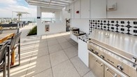 This rooftop terrace is a well-appointed outdoor amenity space designed for elevated communal living. The design aesthetic is immediately striking, anchored by bold black-and-white geometric patterned tilework that adorns both the countertops and backsplashes of the outdoor kitchen area. The outdoor kitchen is fully equipped with stainless steel BBQ grills, a built-in sink, and a water fountain station, making it an ideal setting for social gatherings and al fresco entertaining.

The flooring throughout the terrace consists of large-format speckled stone tiles that lend a clean, modern feel underfoot. A sleek white pergola structure provides partial shade over the dining area, where a substantial teak high-top table surrounded by contemporary metal chairs invites guests to gather and dine outdoors. Patio heaters and additional seating areas are thoughtfully distributed across the space, accommodating a variety of social configurations.

Lush planters with ornamental grasses and small trees add a touch of greenery, softening the urban setting. Glass balustrades along the perimeter ensure unobstructed panoramic views of the surrounding cityscape and skyline, flooding the terrace with natural light throughout the day. The overall ambiance is modern, vibrant, and ideal for both relaxed lounging and lively outdoor entertaining.
