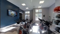 This fitness studio is thoughtfully designed to support an active lifestyle, centered around a spacious spin cycling room that serves as its standout feature. The room is outfitted with a full array of stationary cycling bikes arranged to accommodate group classes, each positioned to face a wall-mounted flat-screen television ideal for instructor-led sessions or on-demand workouts. Large double-hung windows line the walls, flooding the space with abundant natural light and offering views of the surrounding outdoor environment. The ceiling features recessed panel lighting alongside a ceiling fan, ensuring the room remains bright and well-ventilated during intense workouts. The color palette combines a bold navy accent wall with neutral gray tones, creating an energizing yet focused atmosphere. A built-in shelving unit along one wall provides organized storage for fitness accessories such as medicine balls and hand weights. The flooring features a clean, light-toned tile with subtle pattern detailing. A wall-mounted sanitizing station reinforces a commitment to cleanliness and user well-being. Overall, the space strikes a balance between functional design and an inviting ambiance, making it well-suited for both individual training and structured group fitness programming.