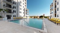 This outdoor amenity space is defined by its generously sized swimming pool, set against the striking white facade of a modern high-rise tower. The pool deck is finished in light-toned tile pavers that reflect the abundant Florida sunshine, creating a bright and inviting atmosphere throughout the day. The pool itself features clear, blue water with stainless steel entry railings for easy access.

The surrounding deck is well-appointed with a large selection of sun loungers in bold yellow and navy blue, offering ample space for relaxation. Several large market umbrellas are positioned throughout the deck, providing shaded areas for those seeking relief from direct sun. Tropical plantings and potted palms are integrated into the design, adding a resort-like ambiance to the space.

The pool area benefits from an elevated position, allowing for open views of the surrounding cityscape and tree canopy on the horizon. The high-rise building frames one side of the deck, with multiple balconies visible above, underscoring the scale of the community. The overall design balances contemporary architecture with practical outdoor living, making this pool deck a central social and recreational feature of the property.