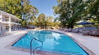 Located at 1515 Hogansville Rd in LaGrange, Georgia, this outdoor amenity space centers around a generously sized swimming pool with a classic geometric shape and clear blue water. The pool deck is finished with a textured, light-toned surface and is lined with multiple lounge chairs and seating areas, providing ample space for relaxation and recreation. A pergola structure sits prominently in the background, offering a shaded gathering spot that adds architectural character to the area. An outdoor grill station is also present, making the space well-suited for casual outdoor entertaining. The entire pool area is enclosed by a wrought iron fence, ensuring both privacy and security. Towering mature trees form a lush green canopy around the perimeter, creating a natural and serene backdrop that enhances the overall ambiance. The surrounding landscaping includes flowering plants and well-maintained grounds that contribute to an inviting, resort-like atmosphere. Abundant natural light fills the space throughout the day, reflecting off the pool's surface and highlighting the vibrant greenery. The combination of functional amenities and natural surroundings makes this outdoor pool area a standout feature of the property in LaGrange, GA.