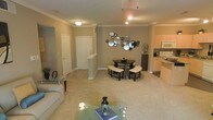 This single-floor residence offers an inviting open-concept layout that effortlessly connects the living, dining, and kitchen areas into one cohesive living space. The living area is finished with plush wall-to-wall carpeting and features crown molding along the ceiling, adding a refined architectural touch. A glass-top coffee table and comfortable seating anchor the space, while decorative accents contribute to a warm, welcoming atmosphere. The dining area is highlighted by a compact, elegantly styled table with matching benches and stools, perfectly suited for intimate gatherings. An eye-catching arrangement of circular mirrors adorns the accent wall, serving as a striking focal point that enhances the room's depth and character. Track lighting overhead provides targeted illumination throughout the open space. Adjacent to the dining area, the kitchen is visible and accessible, equipped with standard appliances including a microwave and refrigerator, and features light-toned cabinetry with a convenient counter overhang. A tiled entryway near the front door provides a clean transition into the carpeted living areas, with decorative columns framing the passage. The overall ambiance is neutral and modern, with warm beige tones creating a cohesive and comfortable interior.