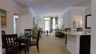 This single-floor residence is thoughtfully designed with an open layout that seamlessly connects the primary living spaces. Crown molding runs throughout, adding a refined architectural detail that elevates the overall aesthetic. Neutral tones on the walls and plush carpeting in the main living areas create a warm, cohesive atmosphere.

The living room is a comfortable and well-lit space, benefiting from a glass door and large windows dressed with floor-length drapes that allow natural light to filter in while offering views of the outdoors. A circular glass coffee table anchors the seating arrangement, complemented by decorative accents including a striking ornate mirror.

Adjacent to the living room, the dining area accommodates a full dining set and connects directly to the kitchen. The kitchen features a granite-topped breakfast bar with seating, providing an additional casual dining option and a natural transition point between the cooking and living spaces.

The bedroom is a well-proportioned private retreat, while the floor plan also includes a bathroom and additional storage. Overall, the space balances functional layout with tasteful finishes throughout.