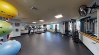This single-floor fitness center is thoughtfully designed to support a comprehensive workout experience. The main exercise area features durable rubber flooring throughout, providing a safe and cushioned surface ideal for a range of physical activities. Recessed panel lighting illuminates the space evenly, while natural light filters in through multiple windows, creating a bright and energizing atmosphere.

The cardio zone is equipped with several treadmills and an elliptical trainer, all from Vision Fitness, arranged in a row along a full-length mirrored wall accented with a wooden ballet-style barre. A wall-mounted television adds entertainment options during cardio sessions.

Strength training equipment includes a multi-function cable machine and a weight bench, alongside a rack of dumbbells for free weight exercises. A dedicated functional training and stretching area features stability balls in multiple sizes, medicine balls, exercise mats, and balance tools organized on storage racks. A wall-mounted fan aids ventilation, and a hand sanitizing station promotes hygiene throughout the facility. Informational signage is posted throughout the space, and a scale is available for health tracking. The light blue painted walls contribute to a calm, motivating environment suited for daily fitness routines.