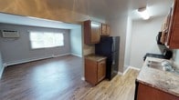 This single-floor space is designed with an open, functional layout that seamlessly connects the kitchen and living areas. The kitchen features warm medium-toned wood cabinetry with upper and lower storage, dark granite-style countertops, and a stainless steel refrigerator, providing both practicality and a cohesive aesthetic. Wood-look laminate flooring runs throughout the space, lending a clean and contemporary feel underfoot.

The kitchen opens directly into a spacious living area, creating a natural flow ideal for everyday living and entertaining. The living room benefits from a large window dressed with horizontal blinds, allowing ample natural light to fill the room while maintaining privacy. Neutral gray walls complement the flooring and trim throughout, contributing to a calm and understated ambiance.

Crisp white baseboards and trim provide a clean contrast against the neutral tones, and a baseboard heating unit along the living room wall ensures year-round comfort. The overall design is straightforward and well-maintained, offering a move-in-ready space with practical finishes and a bright, welcoming atmosphere throughout.
