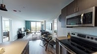 This contemporary single-floor residence offers a well-designed open-concept layout that seamlessly connects the kitchen, dining, and living areas. The kitchen is outfitted with dark espresso cabinetry, light countertops, stainless steel appliances including a range, microwave, and refrigerator, and a classic subway tile backsplash — combining both style and functionality. A glass-top dining table sits adjacent to the kitchen, flowing naturally into the living room, which features light wood-style flooring, a bold teal accent wall, and large sliding glass doors that open onto a private balcony with open outdoor views.

The living room provides a comfortable and stylish retreat, highlighted by a tufted charcoal sofa, vibrant abstract artwork, and an abundance of natural light. Both bedrooms are carpeted and generously sized, each featuring large windows that frame scenic outdoor views. One bedroom showcases a striking green accent wall, adding a pop of color, while the other maintains a calm, neutral palette.

The unit includes two bathrooms. The primary bathroom features a white vanity with a backlit mirror, and a tub-shower combination with marble-style tile. The secondary bathroom offers a glass-enclosed walk-in shower with coordinating tile finishes. A dedicated closet space adjacent to one of the bedrooms adds practical storage to round out this thoughtfully designed residence.