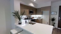 This single-floor residence showcases a thoughtfully designed, modern interior with a strong emphasis on clean lines and contemporary aesthetics. The open-concept kitchen serves as a central highlight, featuring flat-panel cabinetry in warm taupe tones that extend to the ceiling, maximizing both storage and visual impact. Crisp white quartz countertops provide a striking contrast to the cabinetry, while a generous kitchen island with an undermount sink doubles as a casual dining and prep area. A textured stone mosaic backsplash adds depth and character to the cooking zone, complementing the stainless steel appliances, including a built-in microwave and gas range. Recessed lighting throughout the kitchen ensures bright, even illumination, enhancing the clean and polished atmosphere. The open layout creates a seamless flow between the kitchen and adjoining living areas, making the space well-suited for both everyday living and entertaining. A decorative plant on the island counter adds a touch of natural warmth to the otherwise refined, neutral palette. Overall, the space exudes a contemporary and well-appointed character with careful attention to both form and function.