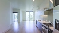 This contemporary single-floor space is defined by its clean lines, open layout, and an abundance of natural light. Floor-to-ceiling windows along the main living area flood the interior with daylight while offering pleasant outdoor views of surrounding greenery, creating a bright and airy atmosphere throughout. A sliding glass door provides direct access to a private balcony, further connecting the interior to the outdoors.

The open-plan design seamlessly integrates the kitchen and living areas, making the space feel expansive and connected. Light hardwood flooring runs throughout, lending warmth and continuity to the minimalist aesthetic. Recessed and track lighting complement the natural light, ensuring the space is well-illuminated at all times.

The kitchen is thoughtfully appointed with modern, two-tone cabinetry — crisp white upper cabinets paired with cool blue-grey lower cabinets — topped with a light stone countertop. A textured tile backsplash adds a subtle decorative element, while a professional-grade stainless steel gas range, an integrated stainless steel sink with a contemporary faucet, and a built-in beverage cooler round out the well-equipped cooking space. The result is a stylish, functional environment designed for modern living.