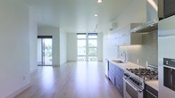 This contemporary single-floor space is defined by its clean lines, open layout, and an abundance of natural light. Floor-to-ceiling windows along the main living area flood the interior with daylight while offering pleasant outdoor views of surrounding greenery, creating a bright and airy atmosphere throughout. A sliding glass door provides direct access to a private balcony, further connecting the interior to the outdoors.

The open-plan design seamlessly integrates the kitchen and living areas, making the space feel expansive and connected. Light hardwood flooring runs throughout, lending warmth and continuity to the minimalist aesthetic. Recessed and track lighting complement the natural light, ensuring the space is well-illuminated at all times.

The kitchen is thoughtfully appointed with modern, two-tone cabinetry — crisp white upper cabinets paired with cool blue-grey lower cabinets — topped with a light stone countertop. A textured tile backsplash adds a subtle decorative element, while a professional-grade stainless steel gas range, an integrated stainless steel sink with a contemporary faucet, and a built-in beverage cooler round out the well-equipped cooking space. The result is a stylish, functional environment designed for modern living.