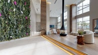 Located at 850 Pacific St in Stamford, Connecticut, this impressive lobby space makes an immediate statement with its dramatic double-height ceilings and thoughtfully curated design. The entry features large-format porcelain tile flooring and glazed entry doors that invite abundant natural light from the streetscape beyond. Towering wood-clad columns rise throughout the space, several of which are adorned with lush vertical living green walls bursting with vibrant foliage and flowering plants, adding a striking biophilic element to the interior. A raised lounge area, accessed via illuminated stepped platforms with warm accent lighting, is anchored by a striking suspended cylindrical fireplace that serves as a bold focal point. This seating zone is finished with wide-plank light oak hardwood flooring and furnished with contemporary neutral sofas and chairs arranged to encourage conversation. Oversized floor-to-ceiling windows on the perimeter frame outdoor views and bathe the lounge in natural light. Sheer white drapery panels soften the structural columns, while organic décor elements such as natural wood side tables and sculptural greenery contribute to the space's warm, nature-inspired aesthetic. The overall ambiance is modern yet welcoming, blending clean architectural lines with rich natural materials.