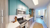 This efficiently designed one-bedroom, one-bathroom unit makes excellent use of its compact footprint, offering a well-organized and modern living environment across a single floor. The galley-style kitchen is a standout feature, showcasing bold teal upper cabinetry paired with warm wood-toned lower cabinets and crisp white quartz countertops. A full suite of stainless steel appliances — including an electric range, over-the-range microwave, dishwasher, and refrigerator — lines the kitchen, while a skylight overhead draws in natural light to brighten the space. The bedroom occupies roughly half the unit's total area and is highlighted by a bank of tall, floor-to-ceiling windows that flood the room with natural light and offer views of surrounding greenery. Light wood-tone flooring runs throughout, contributing to a clean, airy aesthetic. A dedicated open closet nook with a wall-mounted clothing rail provides practical storage within the bedroom area. The bathroom is cleanly appointed with a tub-shower combination, a wood-grain vanity with stone-look countertop, and neutral tile flooring. The unit's modern design, cohesive color palette, and efficient layout make it a well-suited space for comfortable single-occupant living.